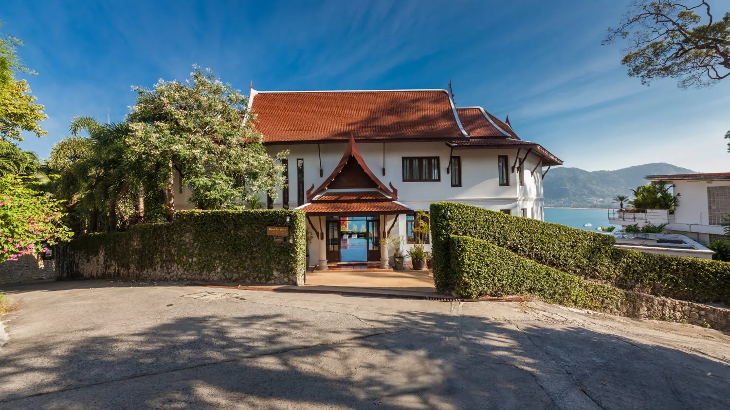 The entrance to Villa Baan Paradise, with its traditional Thai gabled roof and clipped hedge, the bay visible beyond