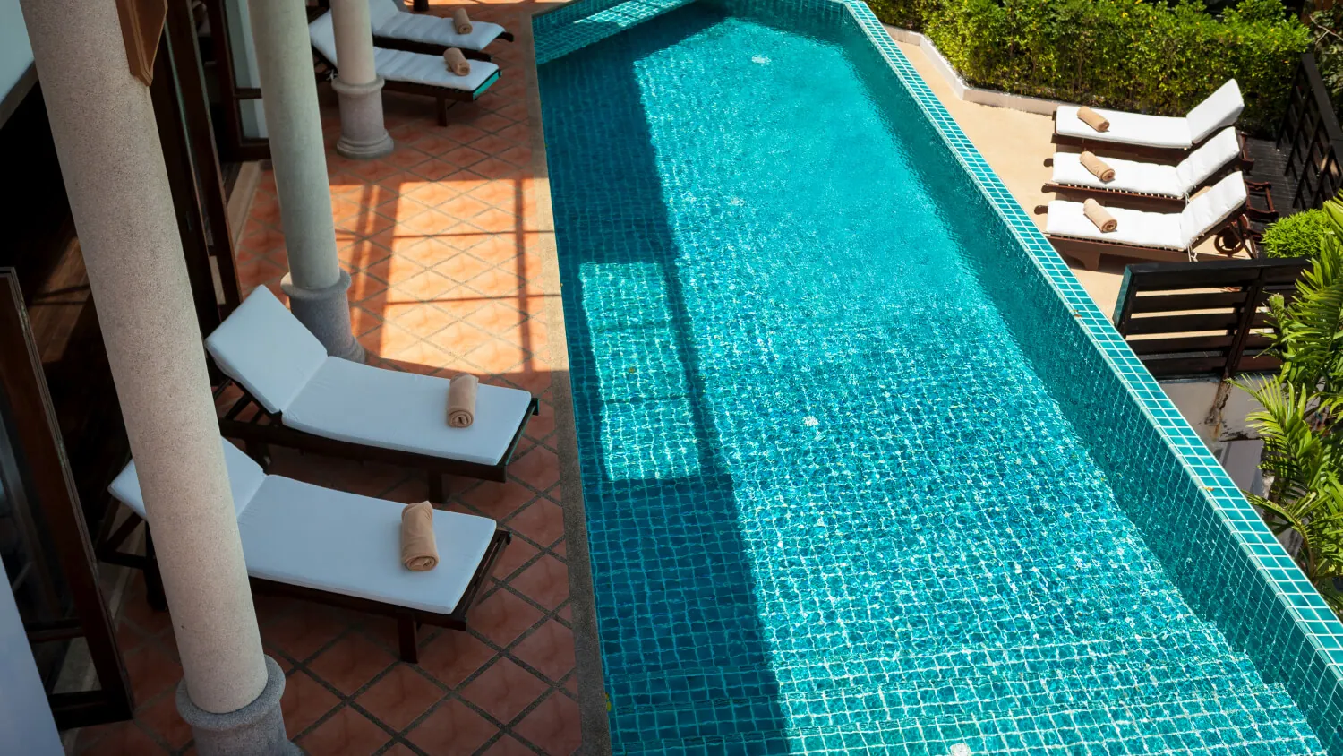 Looking down the turquoise mosaic-tiled infinity pool from the villa colonnade, with loungers on the terracotta deck