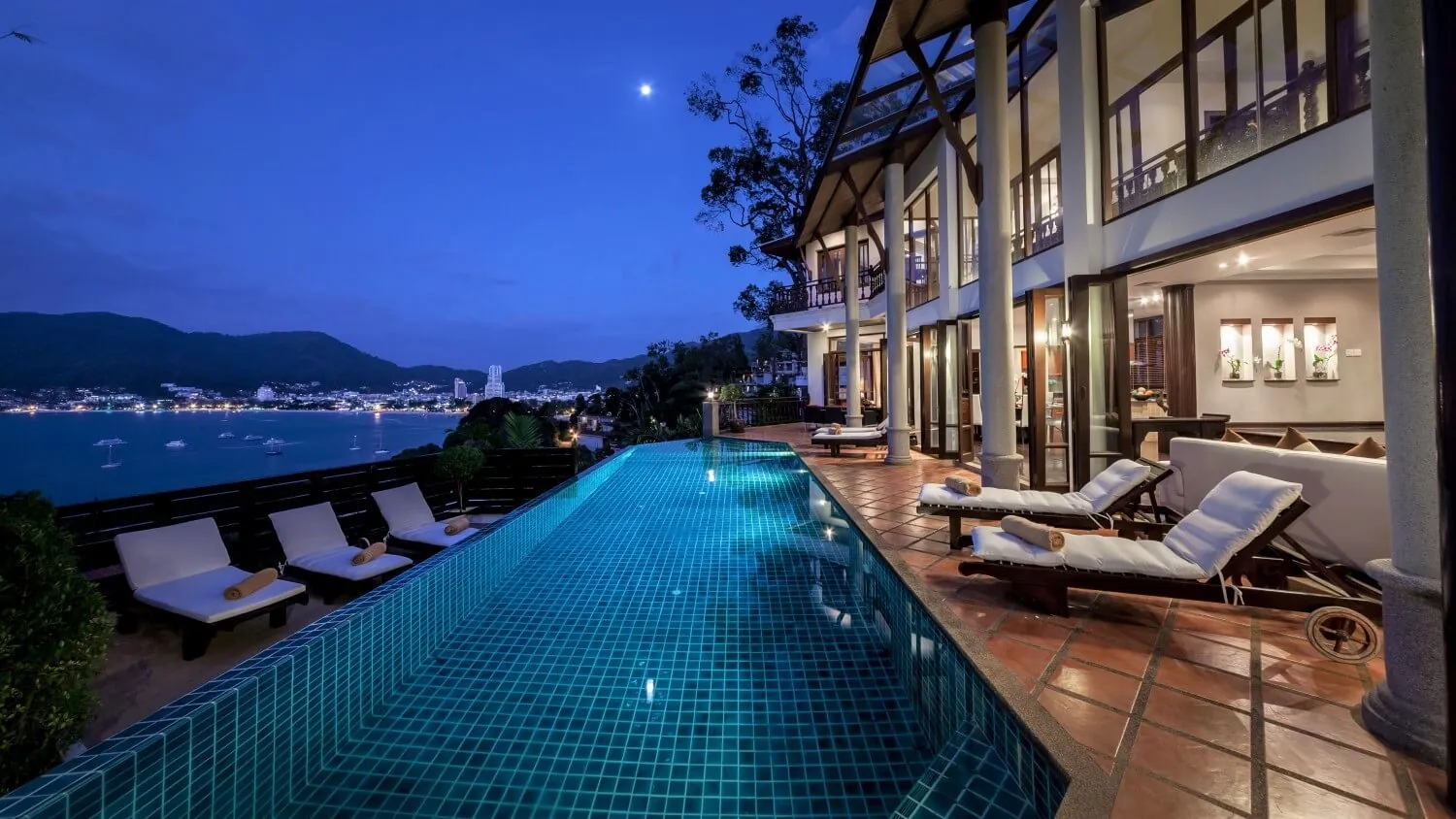 Villa Baan Paradise at blue hour, every room lit, the pool running along the front and the lights of Patong below
